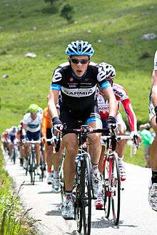 A road racing cyclist in a black and white jersey with blue trim and a conspicuous accented e on each shoulder leading a line of other riders.