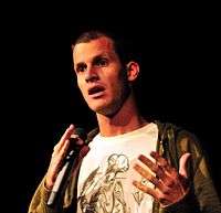 A dark haired Caucasian man wearing a white shirt, and green jacket while performing a stand-up comedy routine