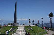 An obelisk on an elevated circular platform, with a few people standing around. Kangchenjunga is visible in the background.