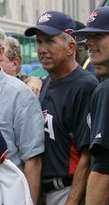 A gray-haired man wearing a navy-blue baseball jersey and cap; he is resting his right hand on the shoulder of an unseen person