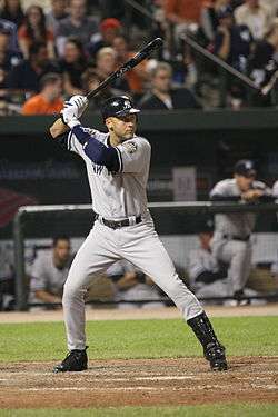 A man in a grey baseball uniform with a navy helmet prepares to swing at a pitch.