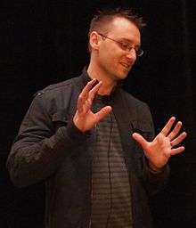 A man with short, spiked up hair stands with his hands up and a microphone clipped to his shirt