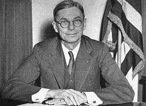 A man poses at a desk. There is an American flag behind him.