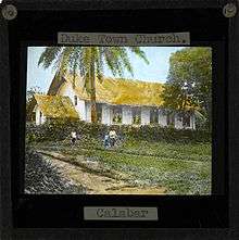 Image of the Duketown Church, Calabar (located within later day Nigeria). Three people stand in front of the white-sided church with a thatched roof. Duketown lies on the Calabar river 50 miles from the coast.