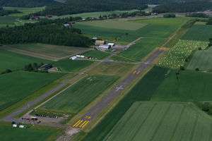 A short, asphalted runway runs though the middle, surrounded by green fields. To the left is a taxiway, and at the top of the runway is a small terminal building