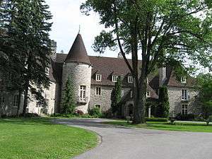 In the foreground is a mowed lawn to the left and a paved road to the right. The road branches to circle an island of grass containing a large tree and a small fountain. A three-storey grey stone building with a brown roof is prominent behind them. It's entrance is partially obscured by the tree, and a three-storey rotunda is clearly visible fronting the right side of the building.