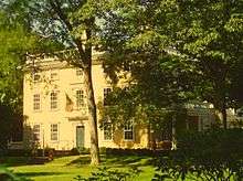 A yellow Federal style house is illuminated by sunlight. It is three stories, with the third story having smaller windows. There is a low railing running around the roof, and there is a covered porch off to the side.