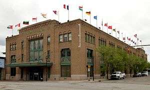 A large, rectangular, tan brick building with green trim and a roof lined with flags