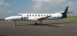 A propellor-driven aircraft with its engines running, coloured white with blue trim, viewed side on and parked on tarmac