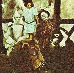 A colour-tinted monochrome photograph of a group of actors. At the rear, a young girl. In front of her, the iron man and scarecrow from 'The Wizard of Oz'; in front of them, people dressed as a lion and another animal.