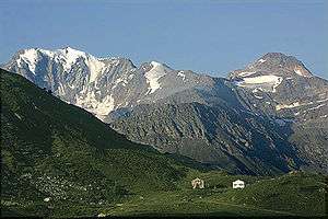 Color photograph of snow-capped mountains in the background with green hills in the foreground