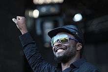 A middle-aged black man wearing a navy blue jacket and baseball cap faces left and holds his clenched right fist in the air above his head.