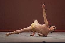 A man, wearing only a dance belt, supporting himself off the ground on one arm and both feet while lifting a fist