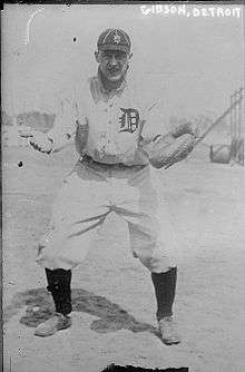 A man in a white baseball uniform with a dark "D" on the front, a dark cap with a white "D", and high socks stands with his arms apart awaiting a throw to his catcher's mitt.