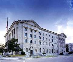 A large white stone building, four stories high, with sections at either end with engaged columns supporting pediments, seen from across the street. On the left side is a colonnade, some small trees and the American flag flying from a pole.