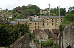 Yellow stone building with grey slate roof and grey chimney, surrounding by houses and trees. In thre foreground is a path with a high stone wall and vegetation.