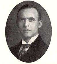 A black and white photograph of a dark haired man in a dark business suit with a white shirt and a tie underneath.