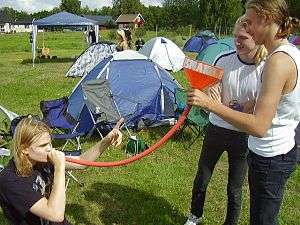 A person sitting on grass outside holds an orange pipe to their mouth while two people standing next to them hold the other end of the pipe with a funnel attached.