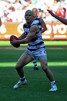 Young male athlete stands in a navy blue and white striped sleeveless shirt and blue navy shorts. With an opponent approaching him, he holds the football to his right side of the body and prepares to dispose of the red football.