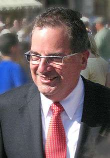 A Caucasian male wearing a black suit with a white dress shirt and red tie is seen among a crowd.