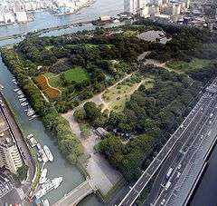 Bird eye view of a park with grassland, trees and ponds surrounded by tall buildings and a big street.