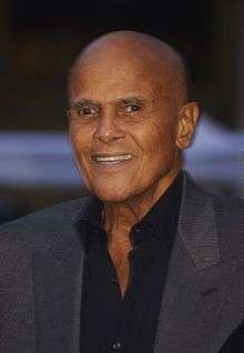 A photo of a bald smiling Jamaican man. He is in his 80s and wearing a dark grey suit over a black collared shirt.