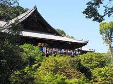 Wooden building with a large hip-and-gable roof built on a hillside. In front of the building there is a wooden railed platform.