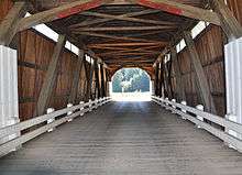 The view is lengthwise along the interior of a covered bridge. Its dark wooden supporting beams, wooden sides and deck, and wooden roof are exposed. Light shines through narrow rectangular openings near its roof. White metal guard rails run the length of the bridge on both sides. A wooded hill lies can be seen beyond the far end of the bridge.