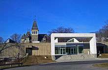 The house with a grey concrete barrier in front of it. To the right is a large square white arch. Behind it is a glass entrance with "Hudson River Museum" written on its right in light gray type.