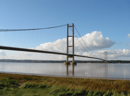  The near pier of a suspension bridge spanning calm blue waters of a wide river estuary.