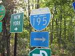 A grouping of road signs, with two fully visible. The left sign is a green square that reads New York right arrow and the right sign is an old shield that reads Interstate 195 right.