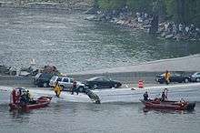 Emergency personnel running on the collapsed bridge and two Minneapolis Fire Department boats in the water with dozens of observers on the bank of the Mississippi
