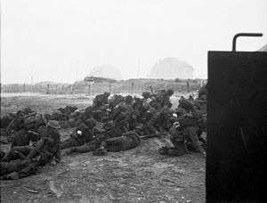 Infantry pile up on the beach. In the distance can be seen barbed wire and a gun emplacement