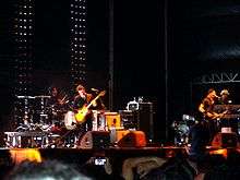 Two guitarists, a drummer and a keyboard player are performing on an orange lit stage in front of an audience. Various pieces of equipment surround the group. The stage features a black backdrop with two strips of vertical lights.