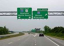 Two green signs are located above a portion of roadway with several vehicles visible on a cloudy day.