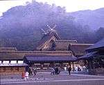 A large wooden building with gabled roof and forked roof finials located beyond other buildings.