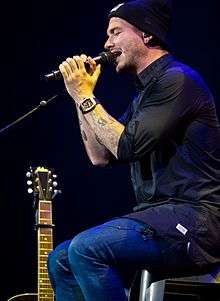 A man sitting on a stool on stage, singing into a microphone.