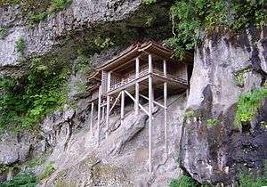 A wooden structure on a cliff face supported by long wooden poles.