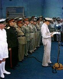 A man in a khaki uniform stands a microphone, reading from papers in his hand. A line of men in smart uniforms stands at attention behind him.