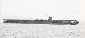 Photograph showing the ocean and a ship at some distance. The ocean has small waves disturbing the surface. The ship's upper deck is nearly flat, and the ship is quite  long compared to the deck's height above the ocean surface. There is a superstructure above the deck that is located somewhat forward of the middle. There is a wake of water at the ship's bow.