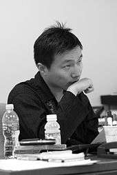 Black-and-white photo of an Asian man seated at a desk, holding his fist to his chin.