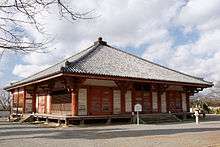 Wooden building with slightly raised floor, open veranda, red beams, white walls and a pyramid shaped roof.