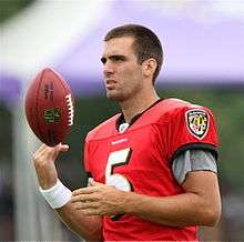 An American football player wearing a red practice jersey (number 5) tossing himself an NFL football while watching a practice.