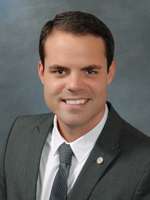 A Caucasian male with thick black hair and wearing a charcoal suit, standing against a grey background