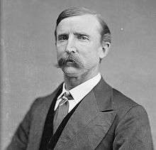 A man with short, dark hair and a mustache wearing a dark jacket and vest, patterned tie, and white shirt