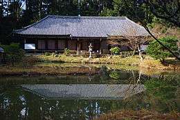 Low and wide building with hipped roof, white walls, beyond a lake.