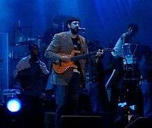 A man holding a guitar with a microphone with pedestal in front of him.