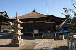 Wooden building with a pyramid shaped roof and an open veranda on the front. A stair leads at one end of the veranda to the building's level.