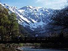 A suspension bridge over a river with high snowcovered mountains in the back.