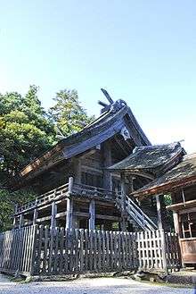 A small wooden building with a veranda and gabled roof raised high above ground. A roofed staircase leads to the gable side of the building.